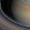 How Old Are Saturn’s Rings? Far Younger Than Saturn — and Slowly Dying Illustration of Saturn tilted with its bright, detailed ring system; the inner ring edge dissolves into faint luminous streams of ice particles drifting toward the planet's hazy gold and blue cloud bands, with subtle stars in deep space.