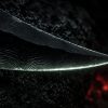 Macro photograph of a black obsidian blade edge showing glassy conchoidal ripples along its sharpened edge, with the dim red glow of lava in the background.