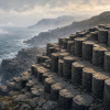 Illustration of a coastal columnar-basalt outcrop: tightly packed dark stone prisms show mostly six-sided but irregular polygonal tops, tall vertically jointed sides and faint transverse growth markings, beside a cold blue sea beneath a misty sky.