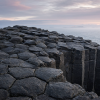 AI-generated geological illustration of a coastal basalt outcrop, showing adjoining irregular polygonal column tops above tall dark stone pillars, fine horizontal bands on their faces, and a misty sea beyond.