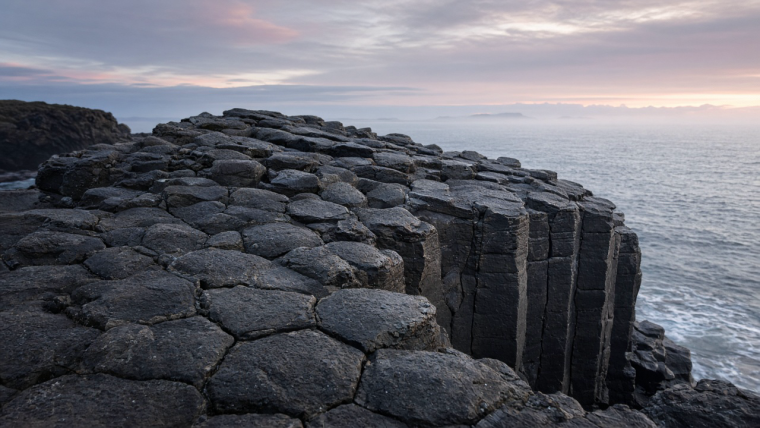 AI-generated geological illustration of a coastal basalt outcrop, showing adjoining irregular polygonal column tops above tall dark stone pillars, fine horizontal bands on their faces, and a misty sea beyond.