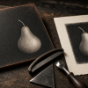 Generated editorial illustration of a mezzotint workshop: a partly burnished copper plate showing a pear, a serrated rocker, scraper and burnisher beside a black-and-white pear print on cream paper. Raking light emphasizes copper texture and the contrast between velvety black and pale highlights.