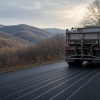 Why Do Road Crews Spray Salt Water Before It Snows? Illustration of a winter maintenance truck applying parallel stripes of salt brine to a clear dark road before snowfall, with bare trees and rolling Appalachian hills under a cold sky.