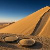 Why Can a Sand Dune Sing More Than One Note? Conceptual illustration of a sunlit golden dune with a steep slip face and a narrow sand-slide channel, with a metal sieve and two trays of sand in the foreground under a clear blue sky; illustrates grain-size experiments, not a photograph of the study site.